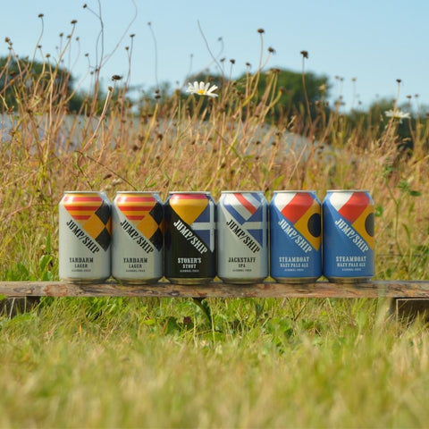 Six cans of Jump Ship Brewing non-alcoholic beer on a wooden plank in a field.