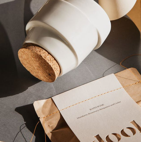 A shoot from the top of white ceramic jar with cork lid laying next to mandarin bag of bath salts. 