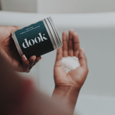 A person holding a tin with bath salts and emptying it on the hand palm. White bath tub and blurred bathroom background.