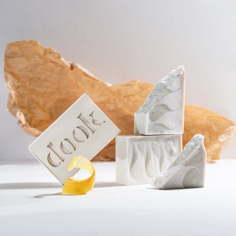 An artistic arrangement of dook handmade bergamot-scented soaps with a pattern of green wavy swirls across a creamy-white bar and one boxed soap with the dook logo, placed next to a lemon peel, the crumpled brown paper behind on the white background.