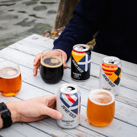 Two people holding glasses of beer and cans on a wooden table outdoors.
