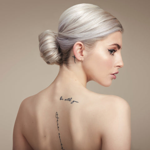 Female model with her back to the camera. Her platinum blonde hair is in a neat low bun, back is bare and she has tattoos down her spine 