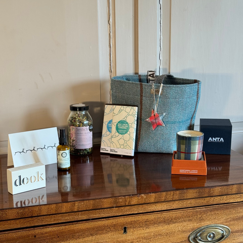 Blue woven Christmas gift bag surrounded by beautiful gifts such as candle, chocolate and soap on a wooden dresser