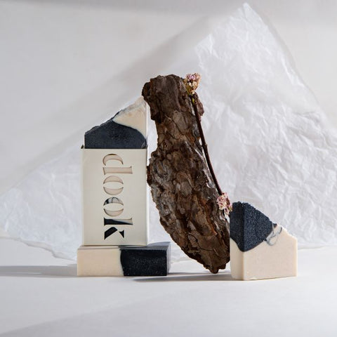 An arty composition of soaps with a piece of wood and dried flowers. A bar of soap lying flat with another, vertically positioned boxed soap on the top and two pieces of a broken soap on the white, crumpled paper background. 