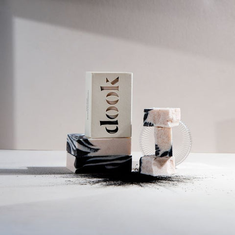 Dook soaps in two piles - as whole and crushed exposing handmade patterns. Sprinkle of a charcoal in the front, white surface and background.