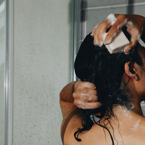 A black haired woman turned back lathering up the shampoo bar under the shower.