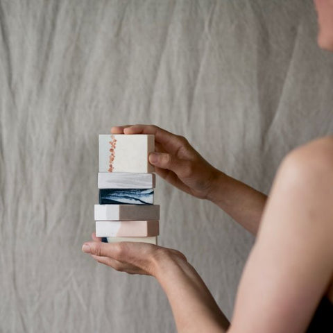 A women holding a stack of six different dook bar soaps on the background of a linen curtain.