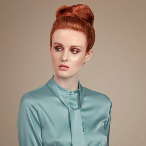 Female model with red hair in a high bun held in place with Moo and Yoo hair spray.  She is facing the camera and looking to her left. She is wearing a pale blue top that is long sleeved and has a high neck. 