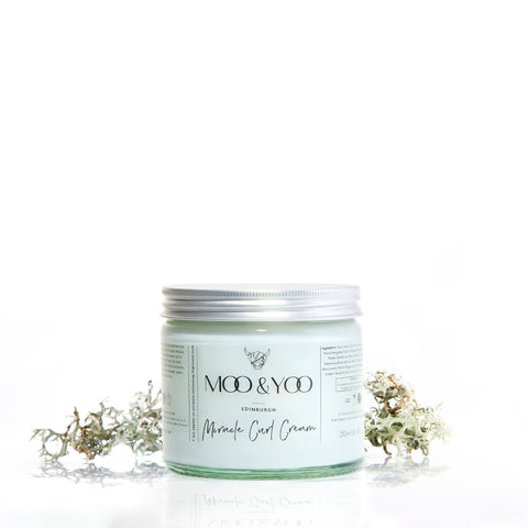 A glass jar of Moo and Yoo Miracle Curl Cream with an aluminium lid on a white background with a sprig of Icelandic moss placed to each side.