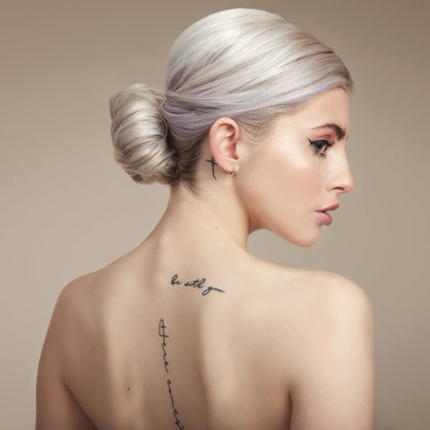 Female model with her back to the camera. Her platinum blonde hair is in a neat low bun, back is bare and she has tattoos down her spine 